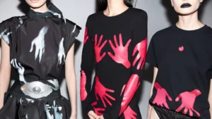 Three models wearing black fashion pieces with bold handprint designs in contrasting colors. The left outfit features white handprints on a dark fabric with a metallic belt, the middle outfit has red handprints across a long-sleeved top, and the right outfit shows a simple black T-shirt with small red handprint accents. The image showcases modern and artistic clothing with a striking visual theme.