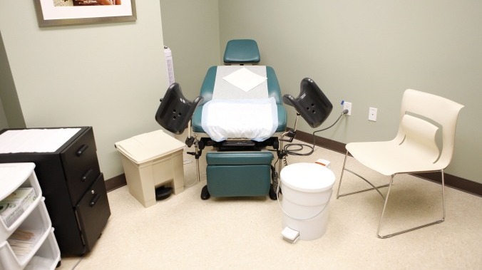 An empty medical examination room featuring a green exam chair with stirrups, covered in disposable paper. The room includes a white chair for visitors, a trash bin, and a small set of drawers holding medical supplies. The setting is clean and minimalistic, with a calm, sterile environment.